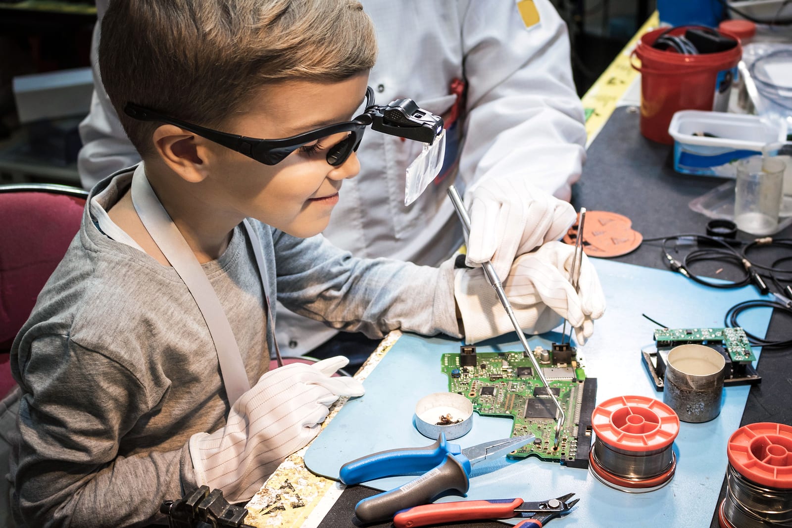 A student wearing safety glasses works on an electronics circuit board with an instructor